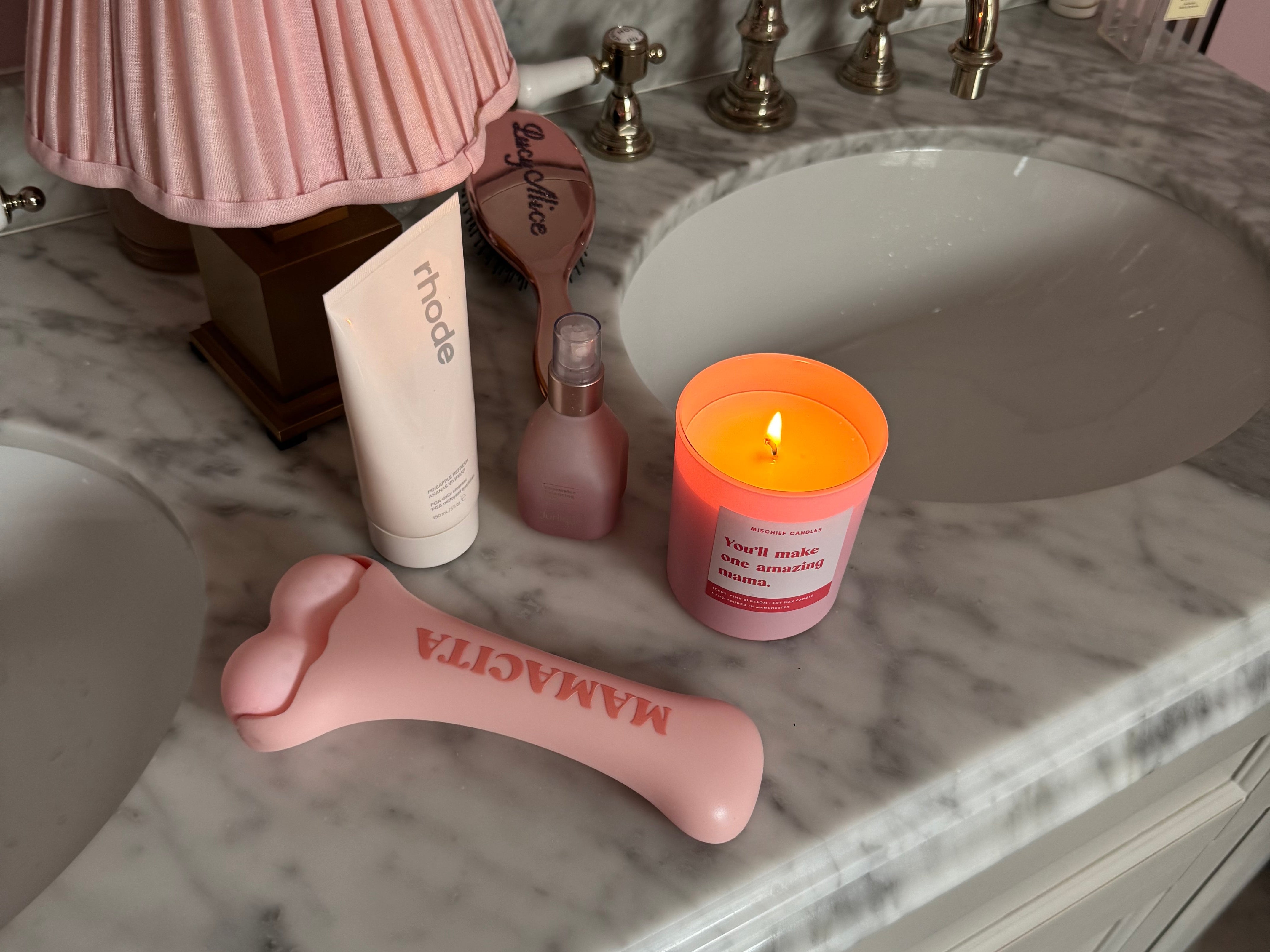A pink silicone prostate massager lies on a white marble bathroom vanity next to a pink pleated lamp, a pink candle, and other toiletries.