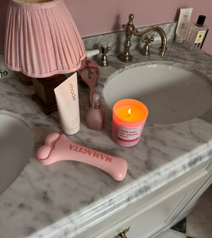 A pink silicone prostate massager lies on a white marble bathroom vanity next to a pink pleated lamp, a pink candle, and other toiletries.