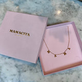 A gold letter necklace spelling 'MAMA' displayed inside a pink presentation box next to its closed lid, which features the brand name 'MAMACITA' in gold lettering, set on a white and gray marble surface.