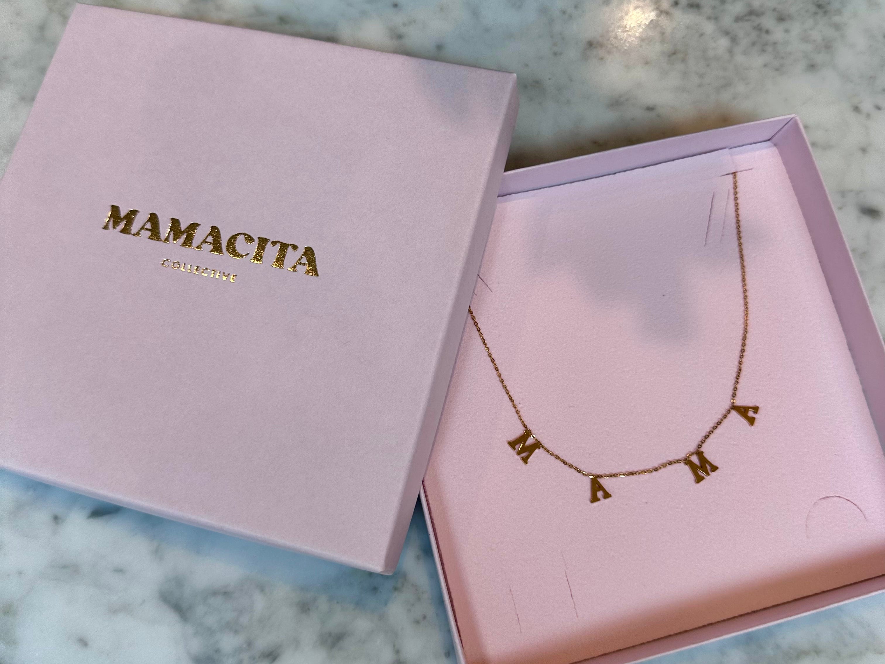 A gold letter necklace spelling 'MAMA' displayed inside a pink presentation box next to its closed lid, which features the brand name 'MAMACITA' in gold lettering, set on a white and gray marble surface.