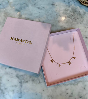A gold letter necklace spelling 'MAMA' displayed inside a pink presentation box next to its closed lid, which features the brand name 'MAMACITA' in gold lettering, set on a white and gray marble surface.