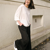 Woman in a white top and black pants standing against a light-colored building.