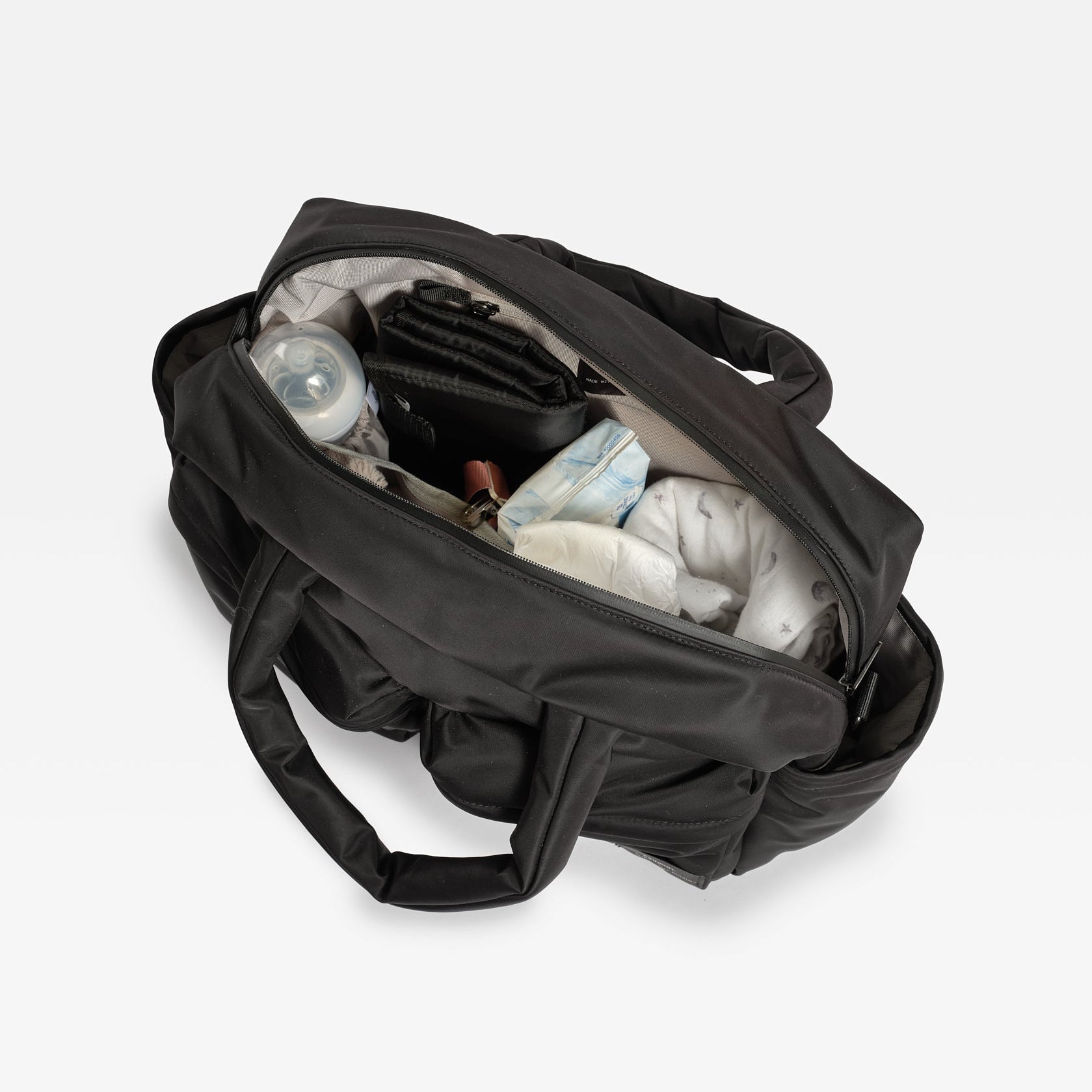A black diaper bag shown open from above, filled with baby items including a bottle, diaper pail, and folded diapers, on a light gray background.