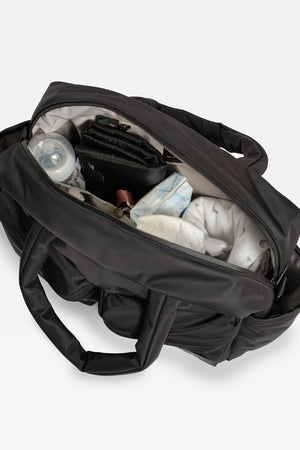 A black diaper bag shown open from above, filled with baby items including a bottle, diaper pail, and folded diapers, on a light gray background.