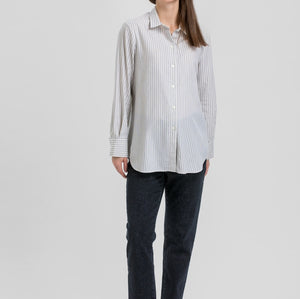 A woman modeling a light gray and white vertically striped button-down shirt, paired with dark charcoal pants and black flats, against a solid light gray background.
