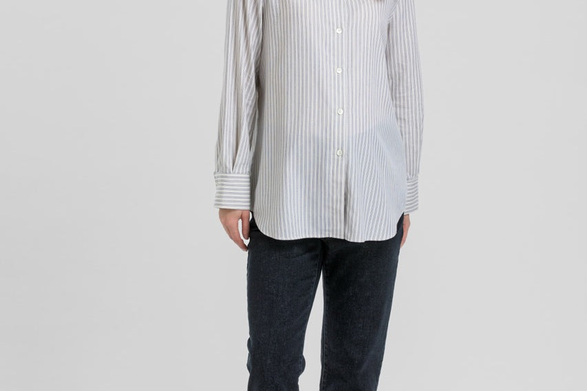 A woman modeling a light gray and white vertically striped button-down shirt, paired with dark charcoal pants and black flats, against a solid light gray background.