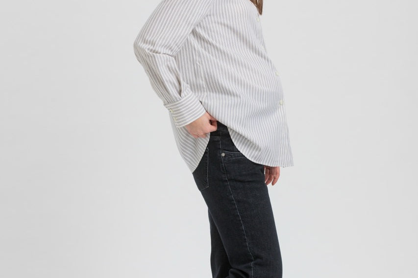 A woman modeling a white long-sleeve button-up shirt with thin vertical black stripes, paired with black jeans and black flat shoes, shown in profile against a light gray studio background.