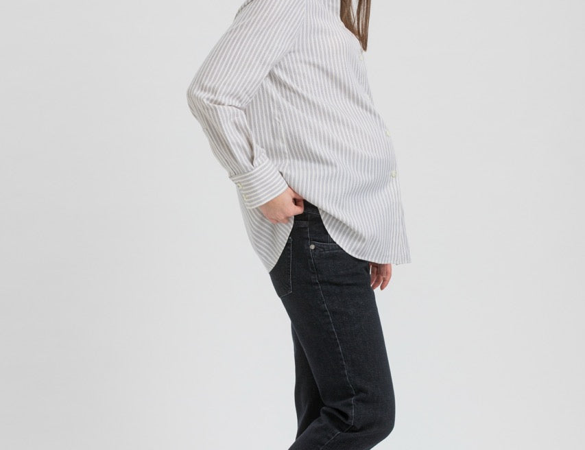 A woman modeling a white long-sleeve button-up shirt with thin vertical black stripes, paired with black jeans and black flat shoes, shown in profile against a light gray studio background.
