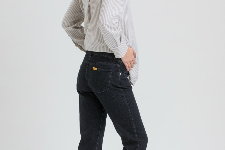 A woman wearing dark wash slim-fit jeans, a light gray and white vertically striped long-sleeve shirt, and black flats, shown from a side-back angle against a light gray studio background.