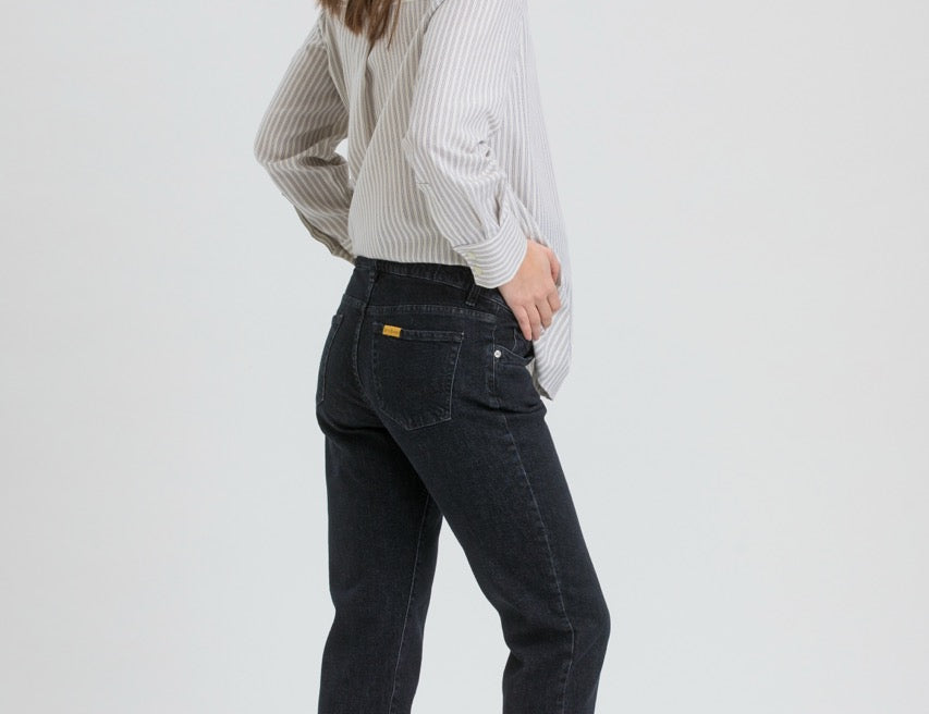 A woman wearing dark wash slim-fit jeans, a light gray and white vertically striped long-sleeve shirt, and black flats, shown from a side-back angle against a light gray studio background.