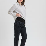 A pregnant woman posing in a studio setting, wearing a white and gray vertically striped long-sleeve button-up shirt, black maternity pants, and black Mary Jane flats.