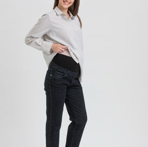 A pregnant woman posing in a studio setting, wearing a white and gray vertically striped long-sleeve button-up shirt, black maternity pants, and black Mary Jane flats.