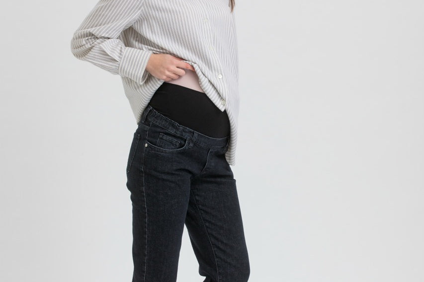 A pregnant woman posing in a studio setting, wearing a white and gray vertically striped long-sleeve button-up shirt, black maternity pants, and black Mary Jane flats.