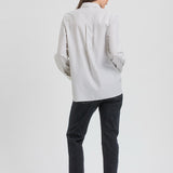 A woman is shown from the back wearing a white long-sleeve button-up shirt with thin vertical gray stripes, paired with dark charcoal pants and black flats, against a plain light gray studio background.
