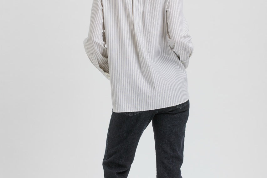 A woman is shown from the back wearing a white long-sleeve button-up shirt with thin vertical gray stripes, paired with dark charcoal pants and black flats, against a plain light gray studio background.