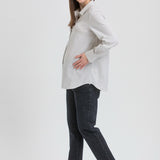 A pregnant woman posing in a light gray and white vertically striped long-sleeve maternity shirt, dark charcoal pants, and black Mary Jane flats against a light gray studio background.