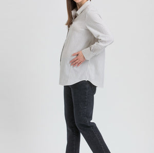 A pregnant woman posing in a light gray and white vertically striped long-sleeve maternity shirt, dark charcoal pants, and black Mary Jane flats against a light gray studio background.