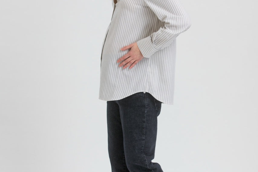 A pregnant woman posing in a light gray and white vertically striped long-sleeve maternity shirt, dark charcoal pants, and black Mary Jane flats against a light gray studio background.