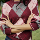 A close-up of a person wearing a chunky knit sweater with an argyle pattern in burgundy, dusty pink, and gray, paired with dark blue jeans, set against a soft-focus outdoor background.