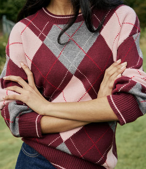 A close-up of a person wearing a chunky knit sweater with an argyle pattern in burgundy, dusty pink, and gray, paired with dark blue jeans, set against a soft-focus outdoor background.