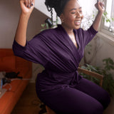 Woman in a purple dress sitting on a chair with her arms raised, smiling.