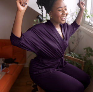 Woman in a purple dress sitting on a chair with her arms raised, smiling.