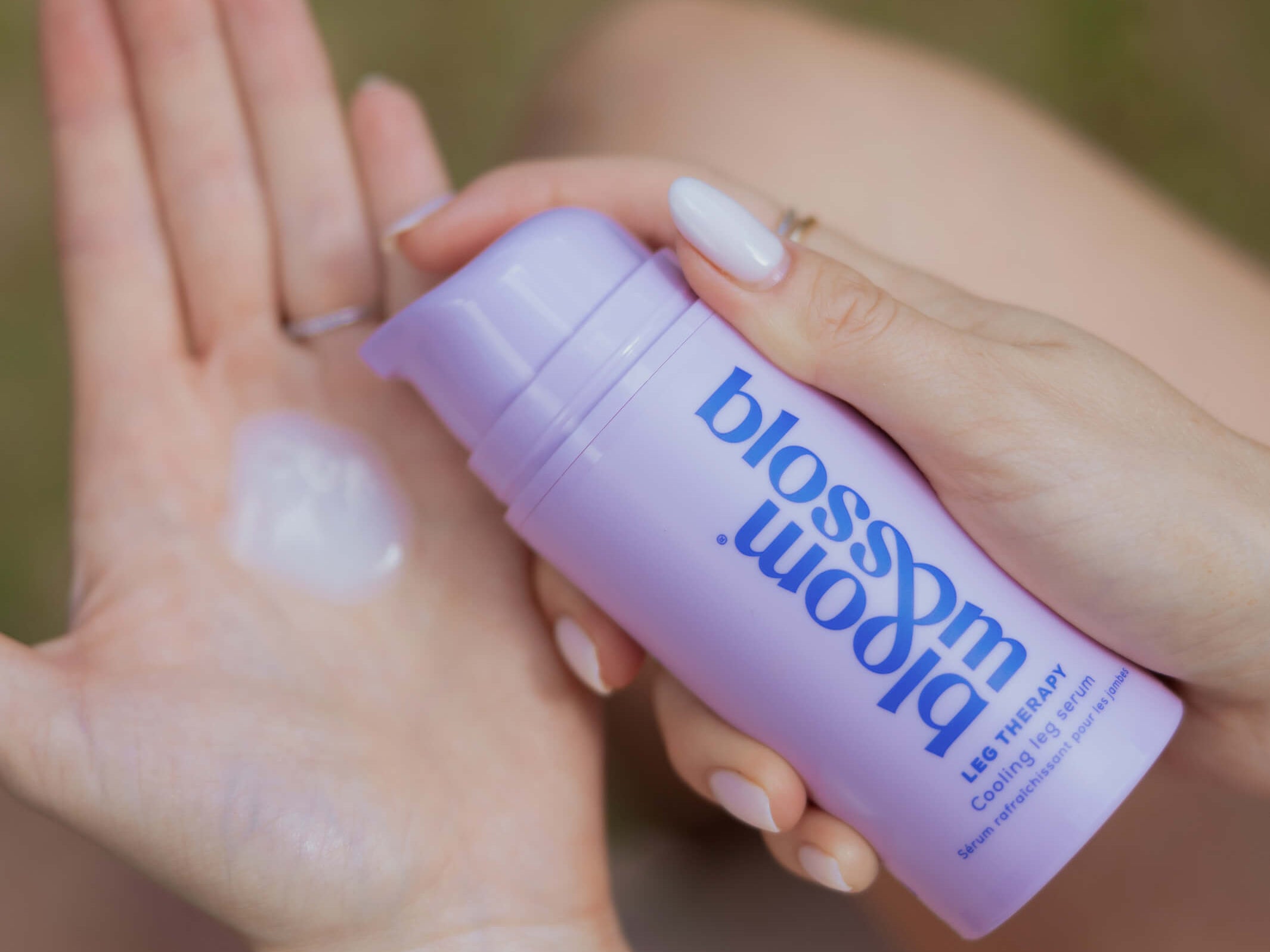 A close-up of a person applying a translucent cream from a lavender pump bottle labeled 'bloom blossom' onto their palm, with a blurred outdoor background.