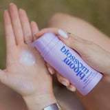 A close-up of a person applying a translucent cream from a lavender pump bottle labeled 'bloom blossom' onto their palm, with a blurred outdoor background.