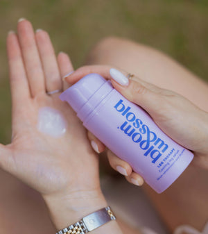A close-up of a person applying a translucent cream from a lavender pump bottle labeled 'bloom blossom' onto their palm, with a blurred outdoor background.