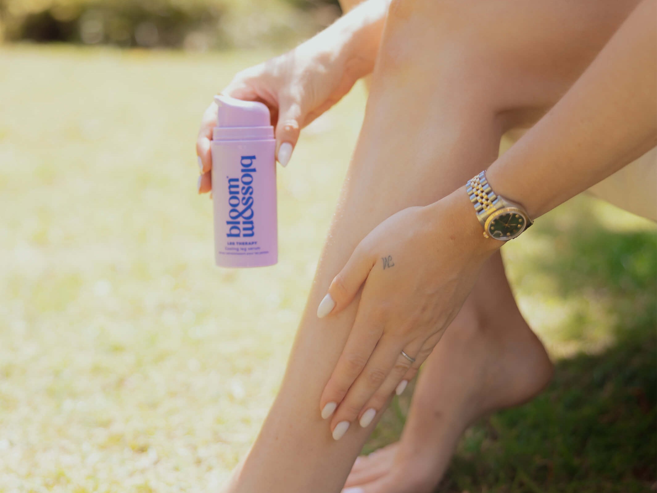 A woman outdoors on grass holds a pink bottle labeled 'blossom balm' while applying a pink product to her leg.