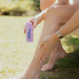 A woman outdoors on grass holds a pink bottle labeled 'blossom balm' while applying a pink product to her leg.