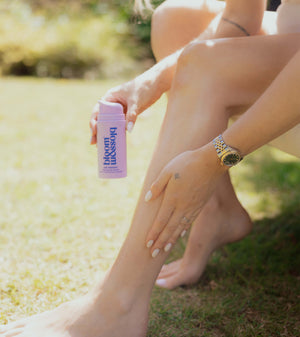 A woman outdoors on grass holds a pink bottle labeled 'blossom balm' while applying a pink product to her leg.