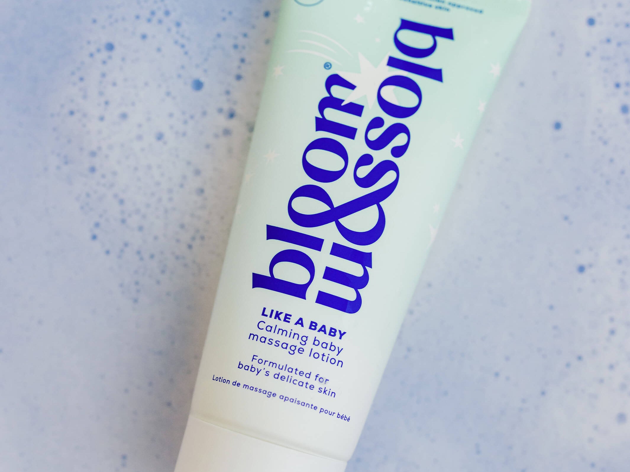A squeeze tube of blossom baby massage lotion on a light blue textured background with small blue speckles. The tube is pale mint green with dark blue text and a white cap.