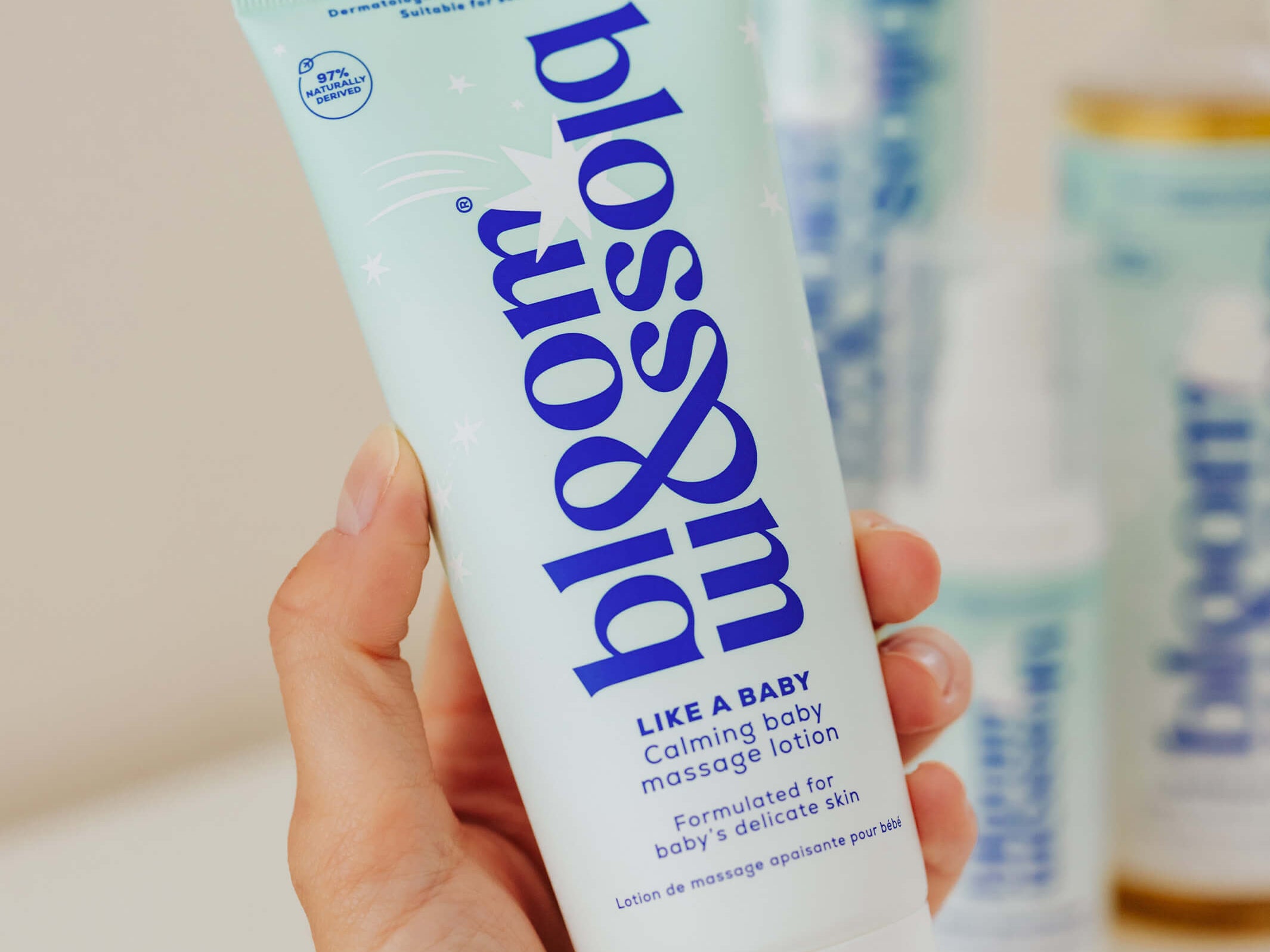 A hand holding a tube of bloom like a baby calming baby massage lotion, with blurred bottles in the background.