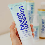 A hand holding a tube of bloom like a baby calming baby massage lotion, with blurred bottles in the background.