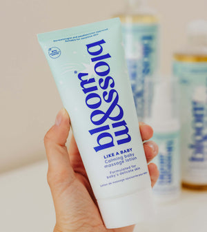 A hand holding a tube of bloom like a baby calming baby massage lotion, with blurred bottles in the background.