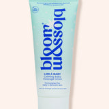 A light blue squeeze tube of bloom & blossom 'Like a Baby' calming baby massage lotion, set against a solid pale peach background.