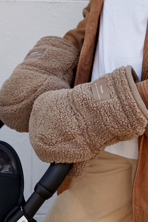 Astrid Eco stroller mittens in Mocha Borg worn by an adult with both hands on a black pram handlebar