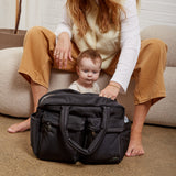 A woman and a baby sit on a beige pouf in a bright room, with a black duffel bag placed in front of them on the carpet.