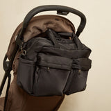 A dark gray diaper bag with multiple zippered pockets and top handles, shown hanging from the handlebar of a brown stroller against a plain beige background.