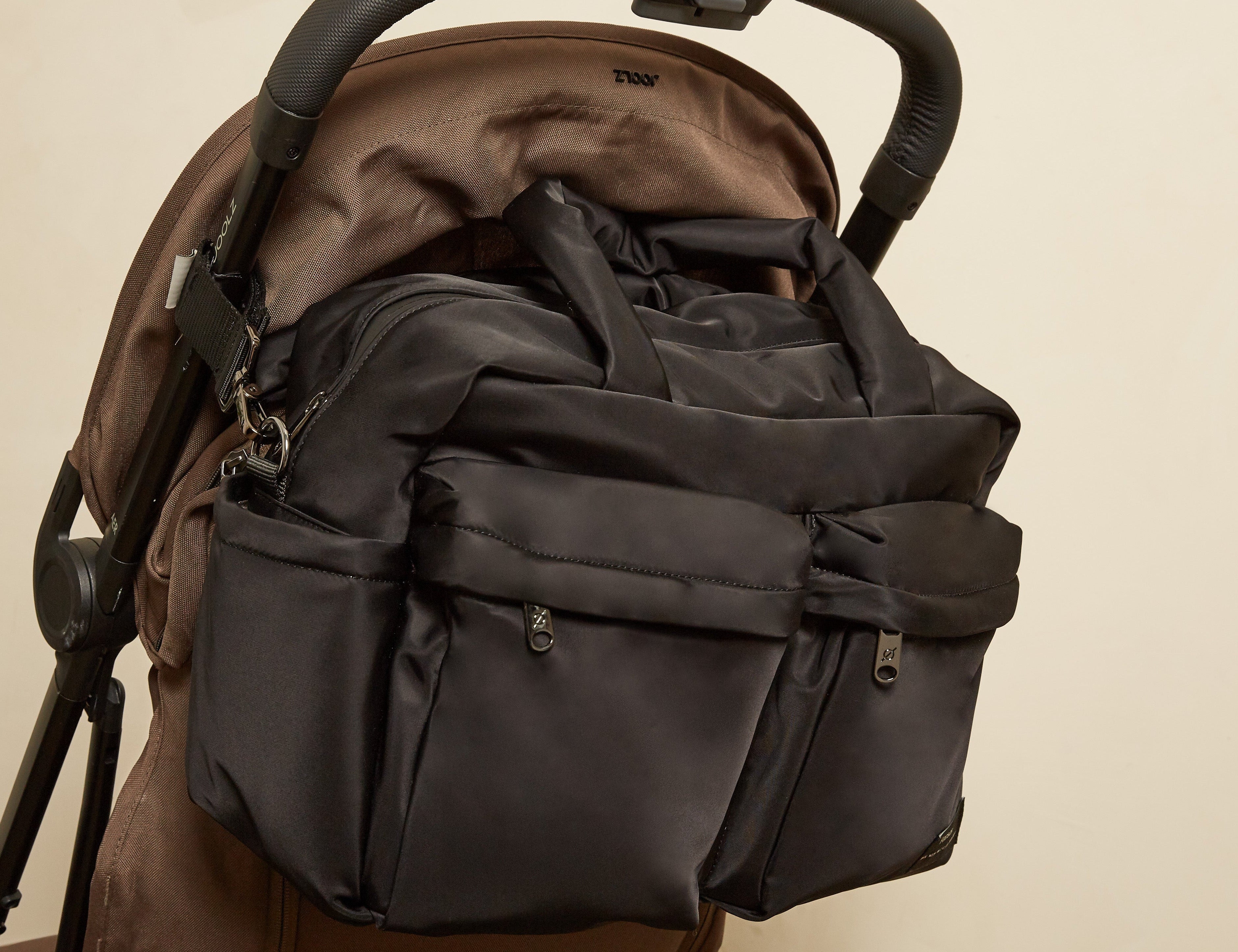 A dark gray diaper bag with multiple zippered pockets and top handles, shown hanging from the handlebar of a brown stroller against a plain beige background.