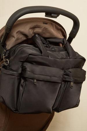 A dark gray diaper bag with multiple zippered pockets and top handles, shown hanging from the handlebar of a brown stroller against a plain beige background.