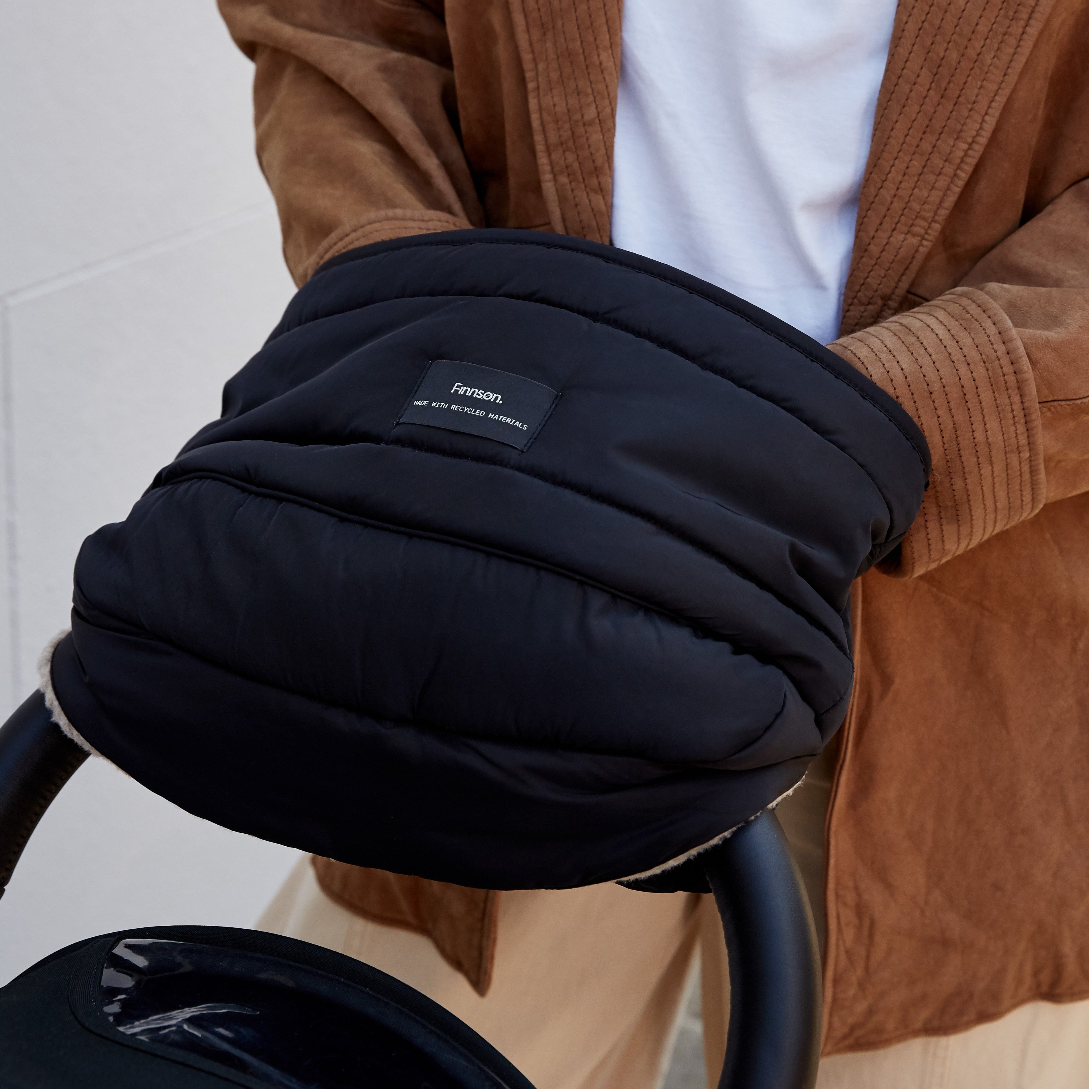 Agnar Eco stroller mitten in black, fitted over a pram handlebar with an adult in a tan jacket resting their hands inside
