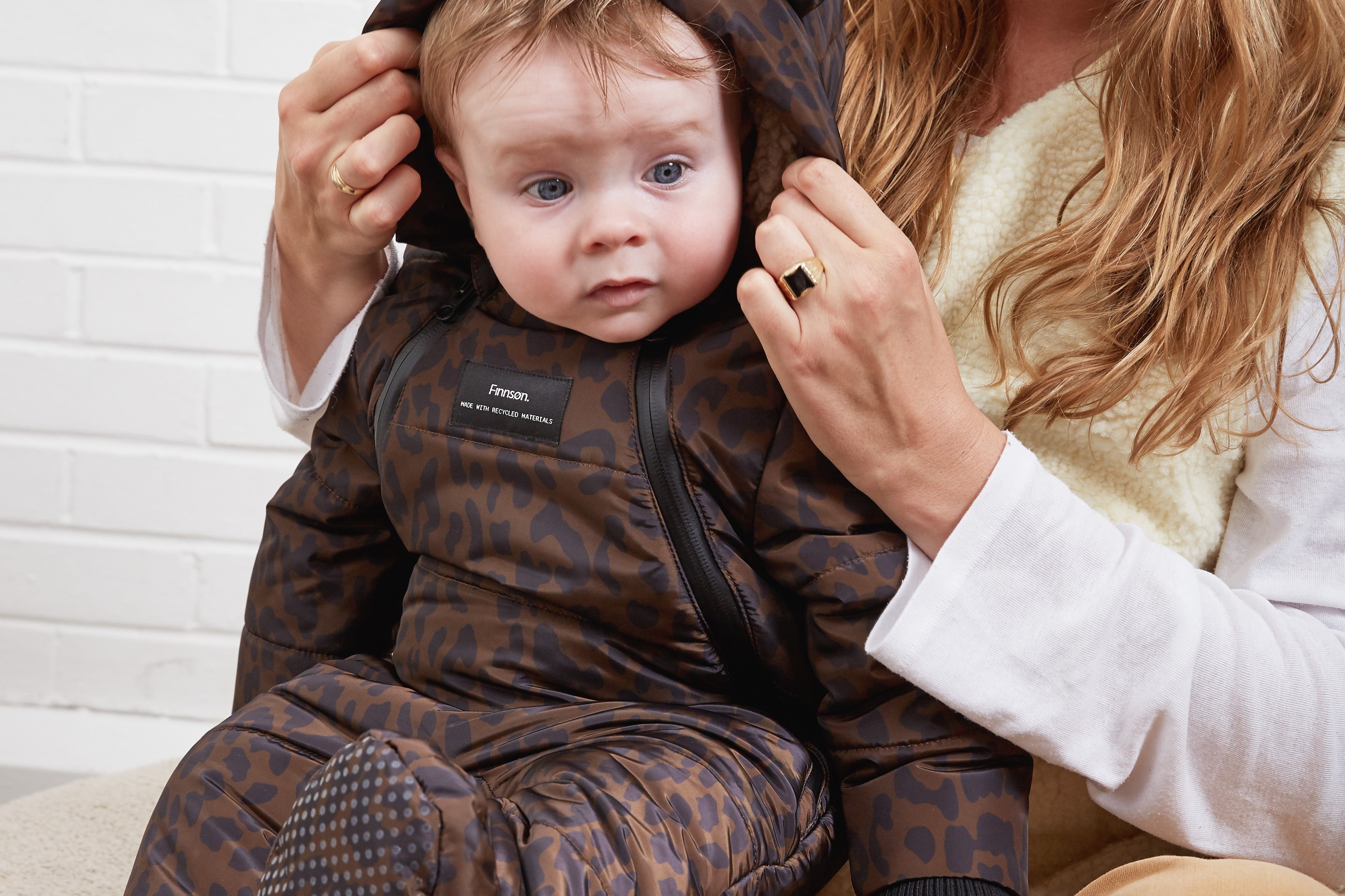 ANDERS Eco Snowsuit Chocolate Leopard worn by a baby sitting on a woman's lap while she adjusts the hood
