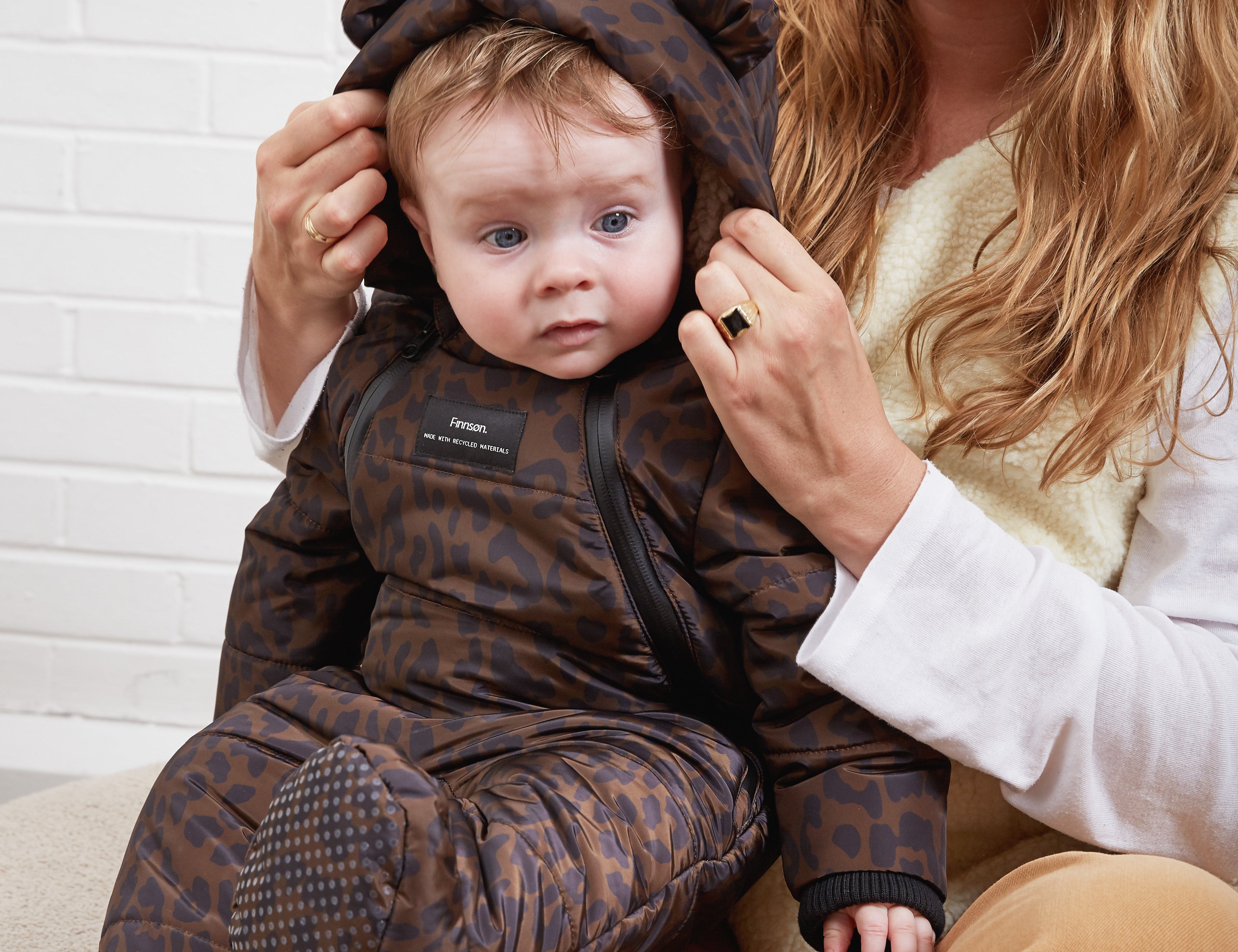 ANDERS Eco Snowsuit Chocolate Leopard worn by a baby sitting on a woman's lap while she adjusts the hood