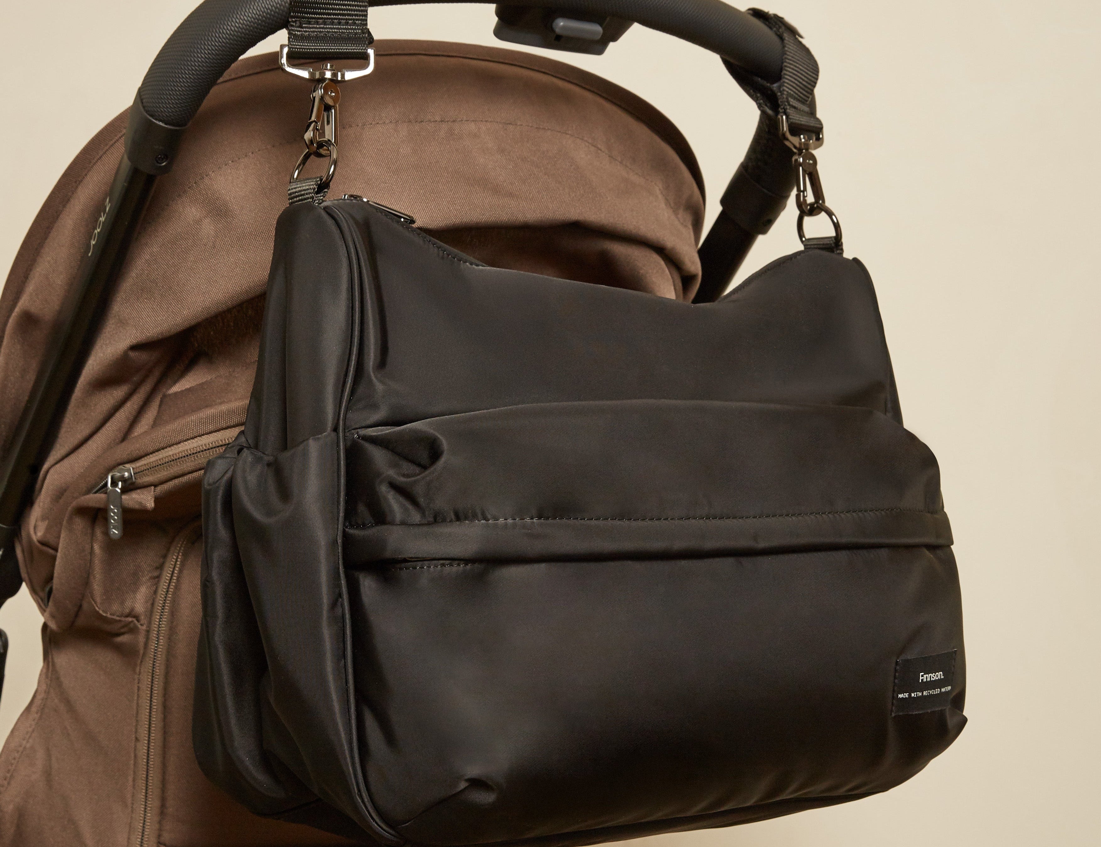 A black diaper bag with a front zippered pocket and side gusset, shown hanging from the handle of a brown stroller against a plain beige wall.