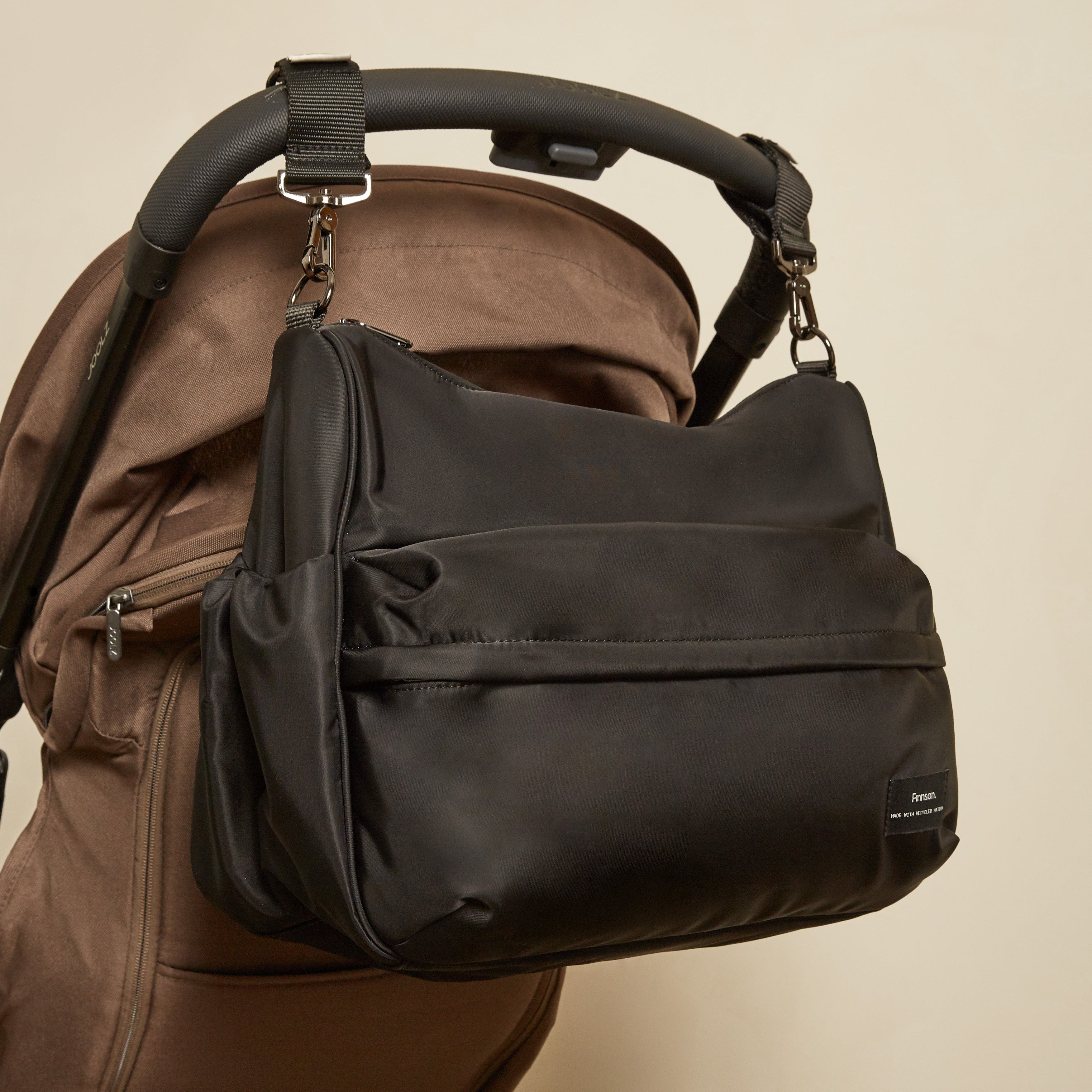 A black diaper bag with a front zippered pocket and side gusset, shown hanging from the handle of a brown stroller against a plain beige wall.