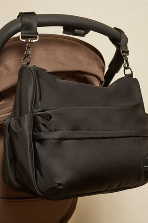 A black diaper bag with a front zippered pocket and side gusset, shown hanging from the handle of a brown stroller against a plain beige wall.