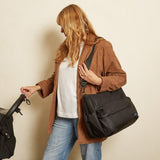 A woman wearing a brown jacket and blue jeans carries a large black messenger bag over her shoulder while walking, with a stroller partially visible on the left against a plain beige background.