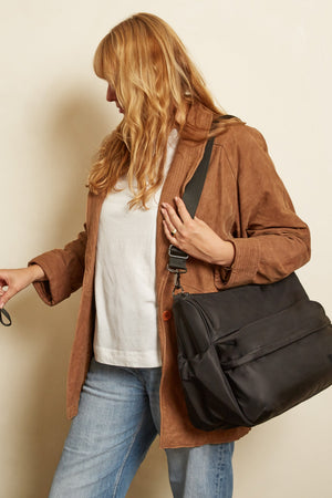 A woman wearing a brown jacket and blue jeans carries a large black messenger bag over her shoulder while walking, with a stroller partially visible on the left against a plain beige background.